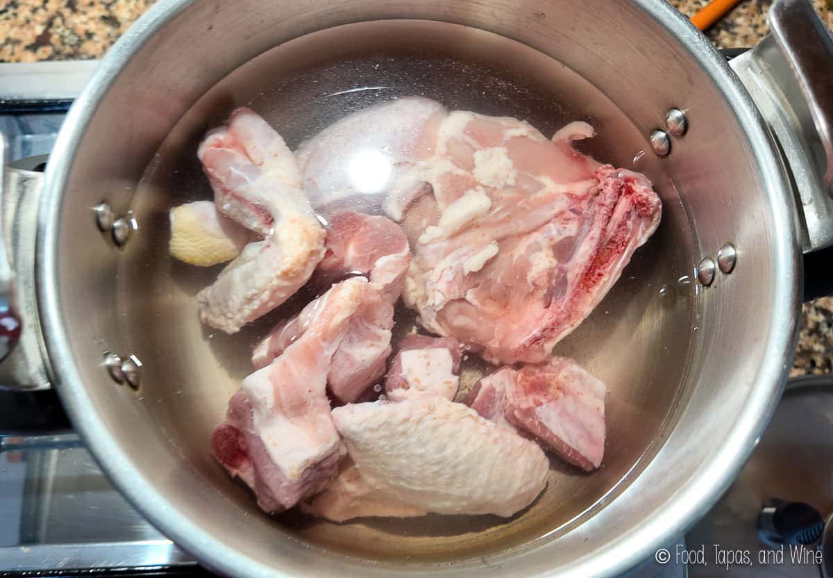 Raw chicken pieces and chunks of pork rib soaking in water inside a metal pot, placed on a stovetop.