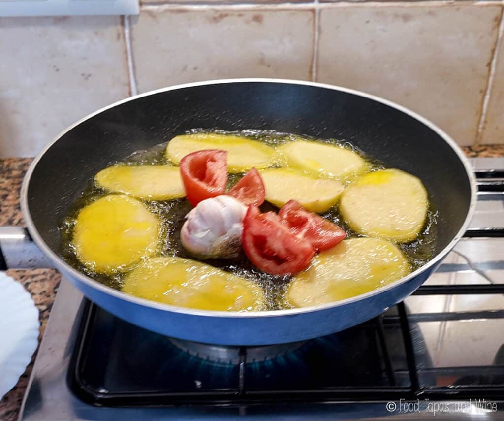 Sliced potatoes, halved tomato, and a whole garlic bulb frying in oil in a black pan on a stovetop.