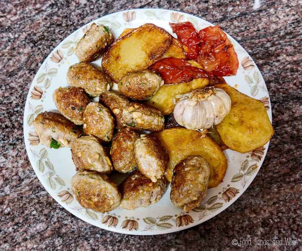 A plate with fried sausage slices, roasted garlic, fried potato slices, and roasted tomato pieces on a granite countertop.
