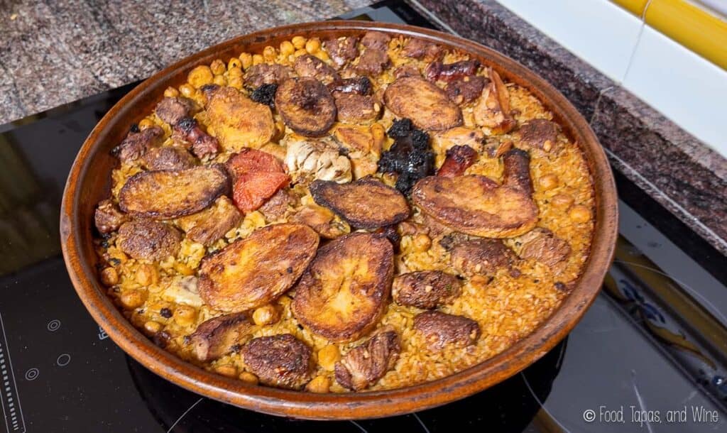 A large round dish filled with baked rice, assorted meats, chickpeas, garlic, and sliced sausages, set on a stovetop.