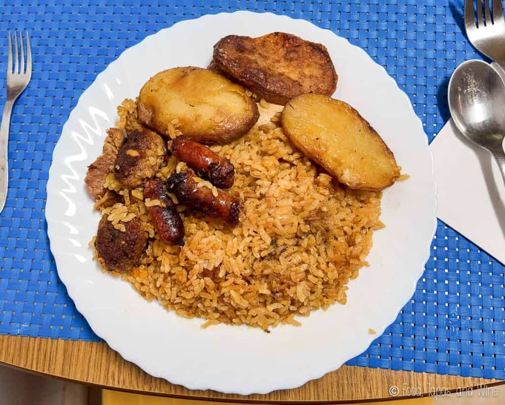 A white plate with seasoned rice, fried potatoes, sausage pieces, and meat, placed on a blue placemat with a fork and spoon on either side.