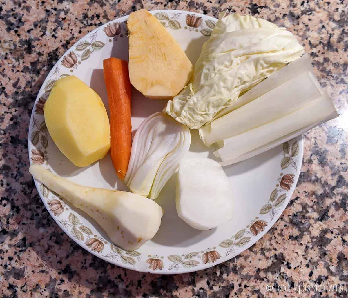 A plate with raw vegetables including potato, carrot, cabbage, leek, parsnip, turnip, onion, and rutabaga on a granite countertop.