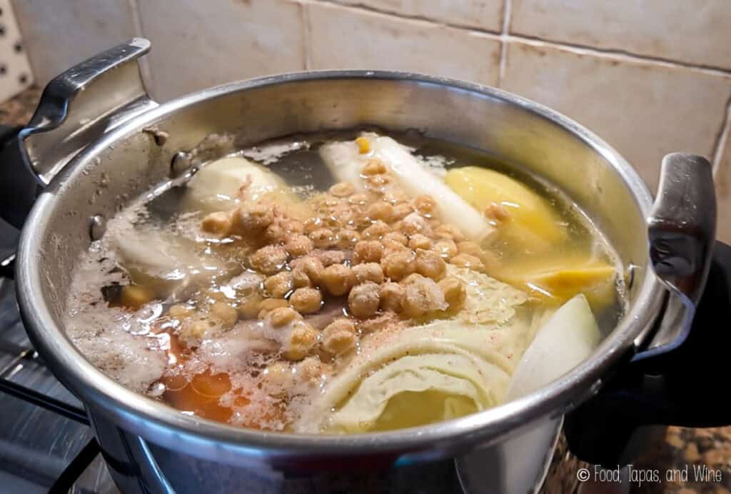 A stainless steel pot on a stovetop contains boiling water with cabbage, chickpeas, potato, and other vegetables.