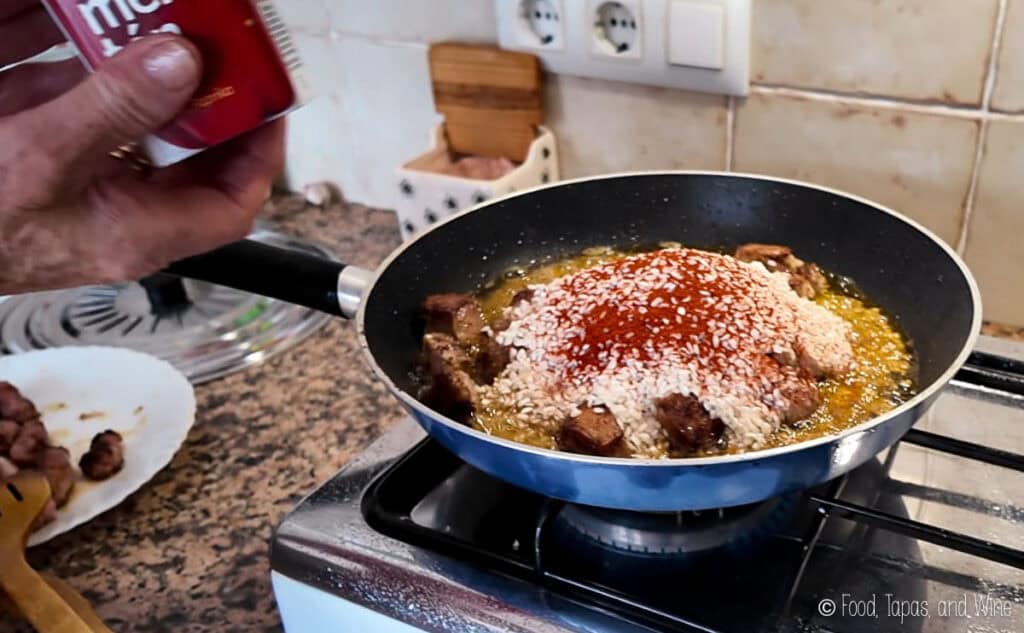 A hand holds a spice container above a frying pan with meat, rice, and paprika cooking in oil on a stovetop.