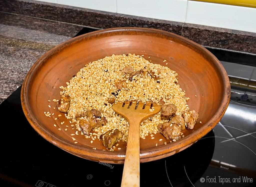 A clay dish with sesame seeds and pieces of dried fruit being stirred by a wooden spatula on an electric stove.