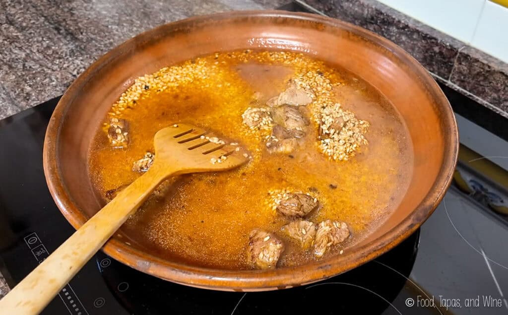 A clay pan with chunks of meat and sesame seeds simmering in a brown sauce on an electric cooktop, with a wooden spatula resting inside.