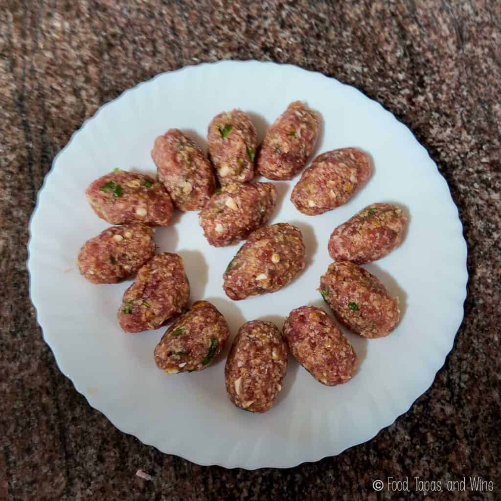 A white plate with fourteen raw, oval-shaped meatballs arranged in a circular pattern on a brown countertop.