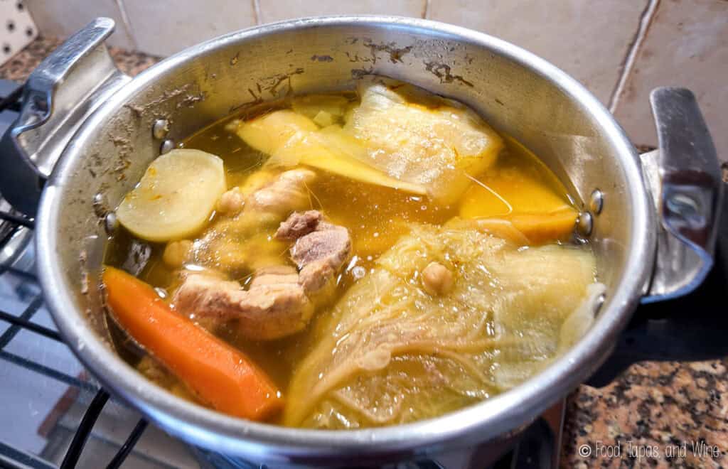 A pot of soup with chunks of meat, carrots, cabbage, chickpeas, and potato simmering on a stove.