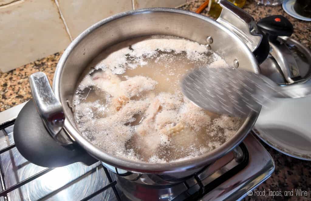 A pot of chicken pieces boiling in water on a stove, with foam being skimmed off the surface using a slotted spoon.