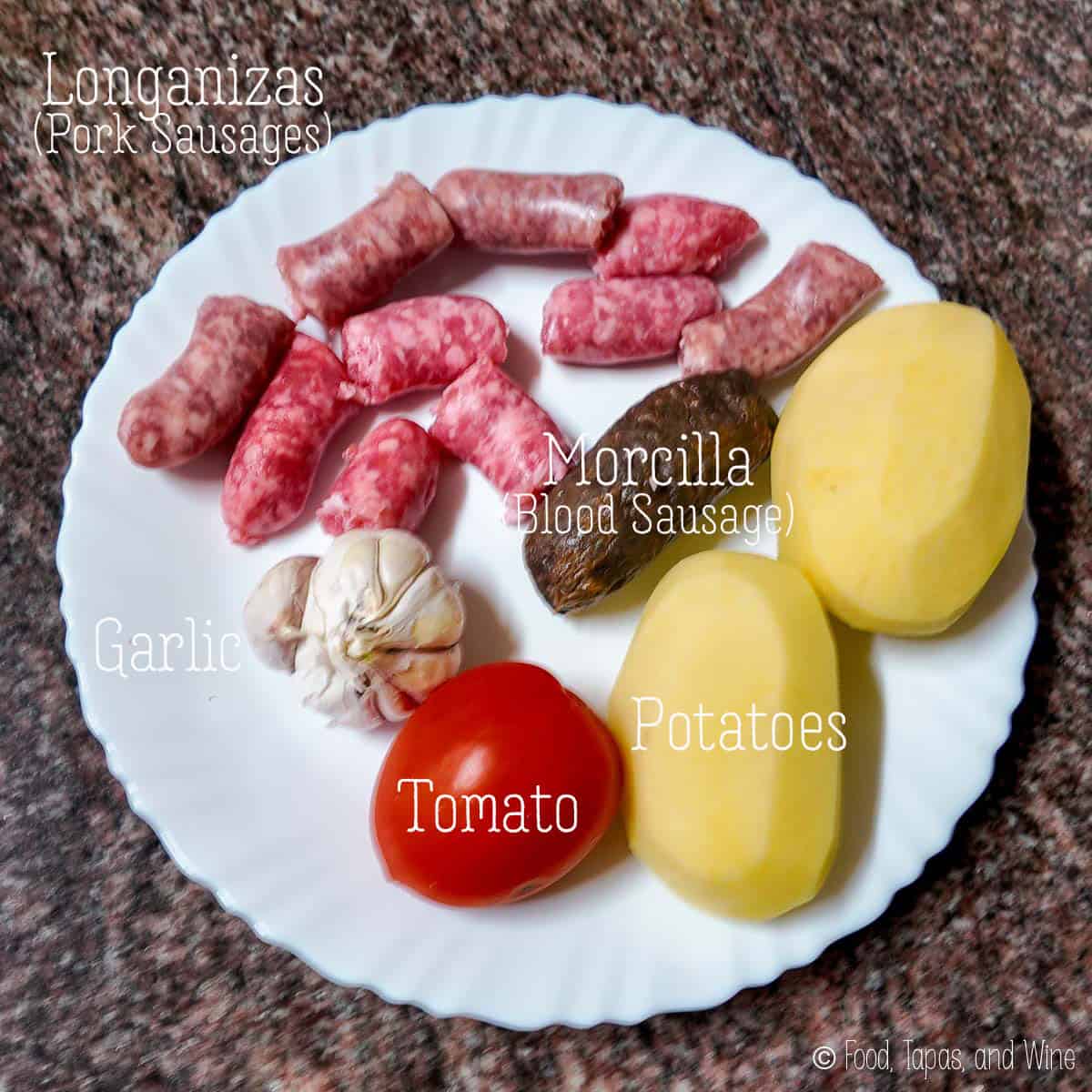 A white plate holds ingredients for arroz al horno: raw pork sausages, blood sausage, two peeled potatoes, a tomato, and a head of garlic, each labeled with text.