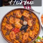 A round dish of Arroz al Horno, featuring rice, chicken pieces, sausage, beans, tomato, and a bulb of garlic, placed on a white table with a side salad.