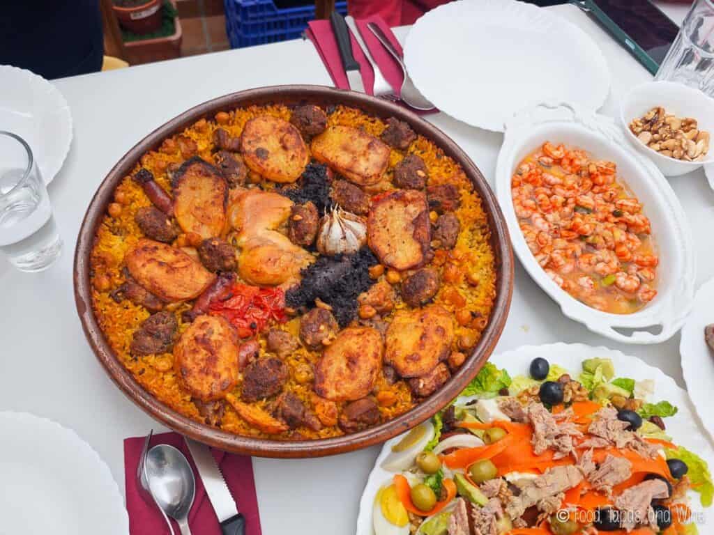 A table set with arroz al horno, a salad with tuna and olives, a shrimp dish, walnuts, and empty plates with cutlery and glasses.