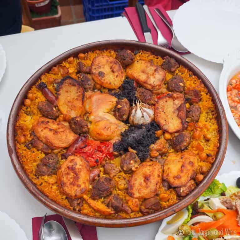 A large dish of arroz al horno with rice, chicken, sausage, potatoes, chickpeas, and a whole garlic bulb, served on a table with plates and cutlery.