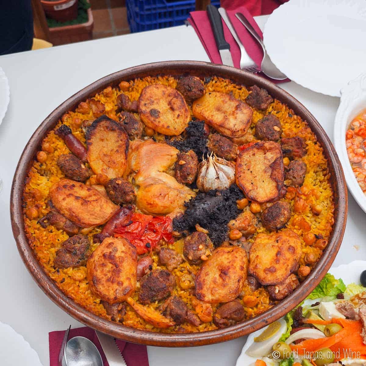 A large dish of arroz al horno with rice, chicken, sausage, potatoes, chickpeas, and a whole garlic bulb, served on a table with plates and cutlery.
